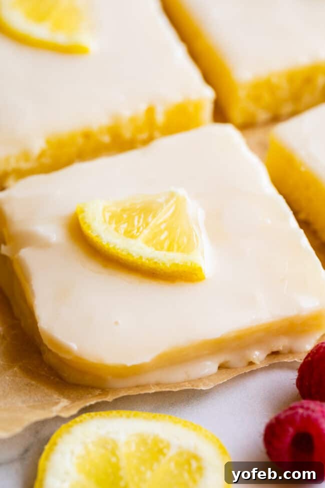Lemon Sheet Cake with Lemon Glaze. This Lemon Sheet Cake with Lemon Glaze is perfect for lemon lovers! A supremely lemony and MOIST cake, paired with a bright lemon glaze. It is perfect for summertime, or anytime! This recipe feeds a crowd and is super easy to make. #lemon #cake #sheetcake #lemoncake #citrus #summer #easy #recipe #zest #tart #withglaze #fromscratch #homemade #glaze #best #Texas #simple #dessert #feedsacrowd #foracrowd squares of lemon sheet cake with glaze, lemon slices, and fresh raspberries on parchment paper.