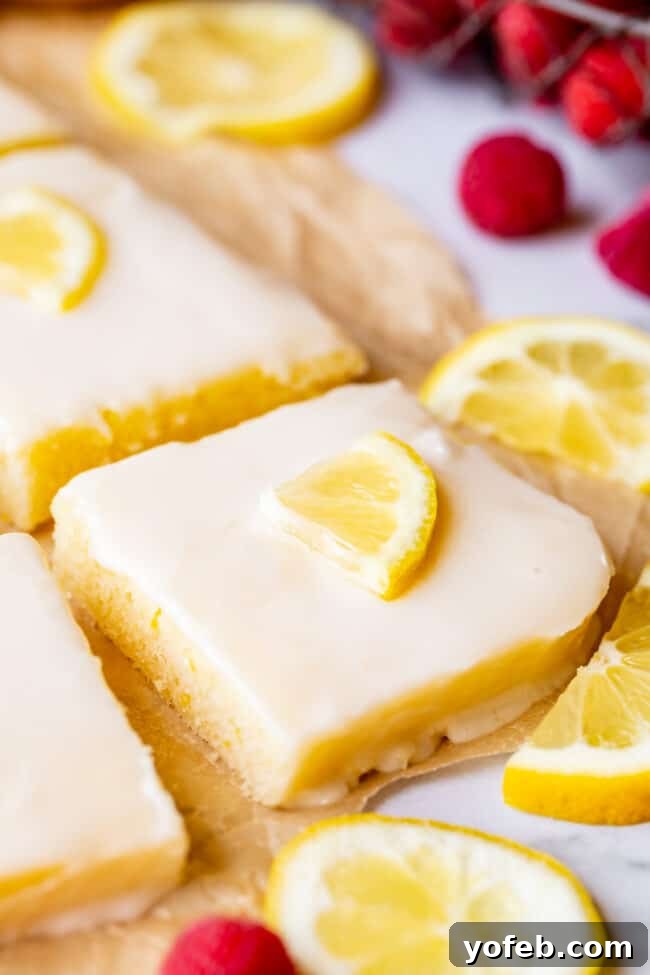 Lemon Sheet Cake with Lemon Glaze. This Lemon Sheet Cake with Lemon Glaze is perfect for lemon lovers! A supremely lemony and MOIST cake, paired with a bright lemon glaze. It is perfect for summertime, or anytime! This recipe feeds a crowd and is super easy to make. #lemon #cake #sheetcake #lemoncake #citrus #summer #easy #recipe #zest #tart #withglaze #fromscratch #homemade #glaze #best #Texas #simple #dessert #feedsacrowd #foracrowd squares of lemon sheet cake with lemon slices and fresh raspberries on a parchment lined baking sheet.