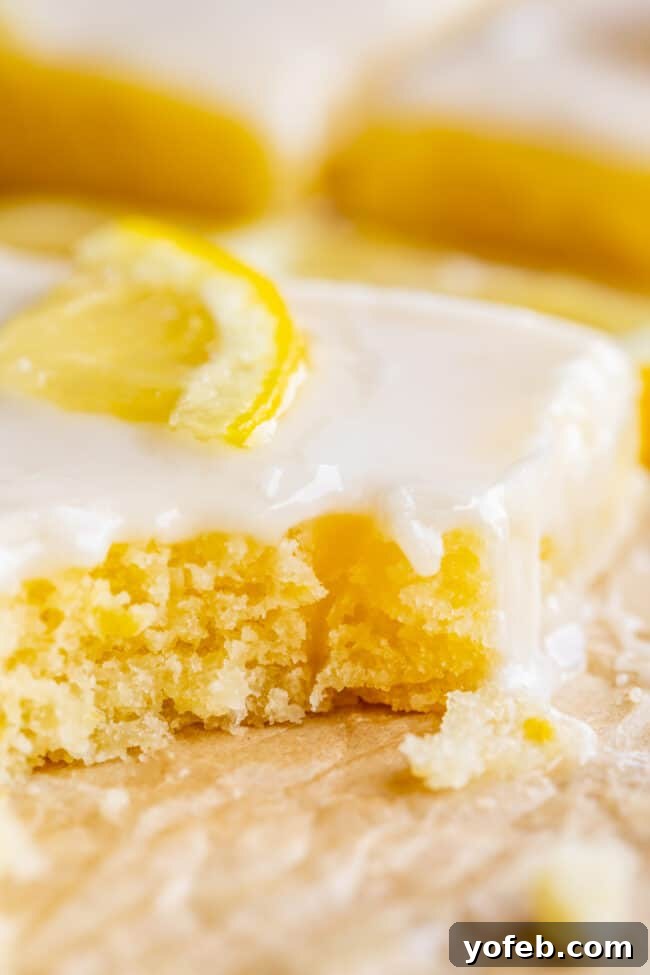 Lemon Sheet Cake with Lemon Glaze. This Lemon Sheet Cake with Lemon Glaze is perfect for lemon lovers! A supremely lemony and MOIST cake, paired with a bright lemon glaze. It is perfect for summertime, or anytime! This recipe feeds a crowd and is super easy to make. #lemon #cake #sheetcake #lemoncake #citrus #summer #easy #recipe #zest #tart #withglaze #fromscratch #homemade #glaze #best #Texas #simple #dessert #feedsacrowd #foracrowd a piece of lemon sheet cake with glaze and a lemon slice with a bite taken out.