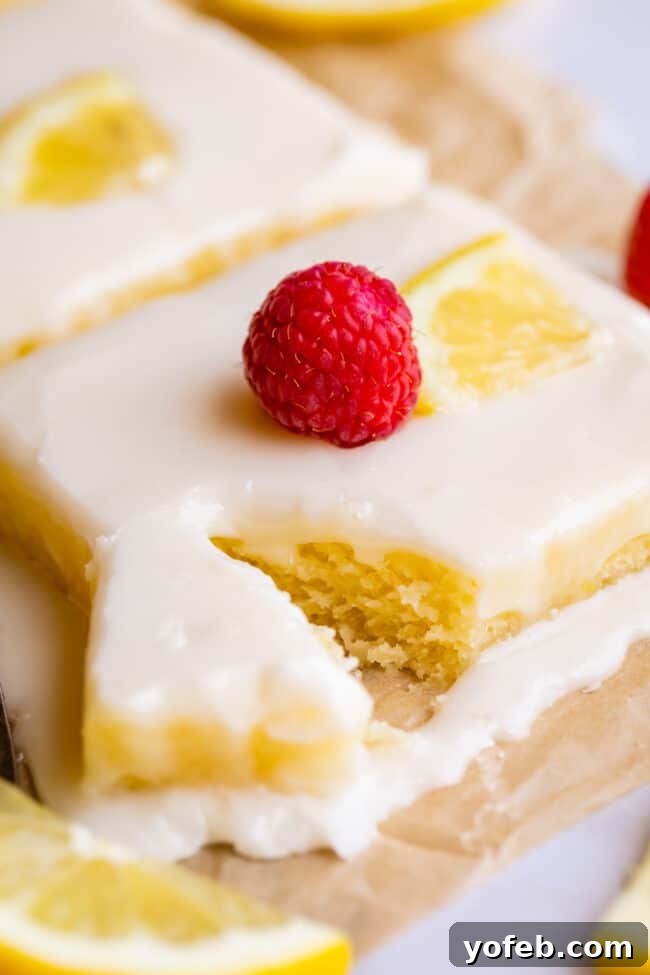 Lemon Sheet Cake with Lemon Glaze. This Lemon Sheet Cake with Lemon Glaze is perfect for lemon lovers! A supremely lemony and MOIST cake, paired with a bright lemon glaze. It is perfect for summertime, or anytime! This recipe feeds a crowd and is super easy to make. #lemon #cake #sheetcake #lemoncake #citrus #summer #easy #recipe #zest #tart #withglaze #fromscratch #homemade #glaze #best #Texas #simple #dessert #feedsacrowd #foracrowd lemon sheet cake with glaze, a sliced lemon, and a fresh raspberry, one bite partially removed.