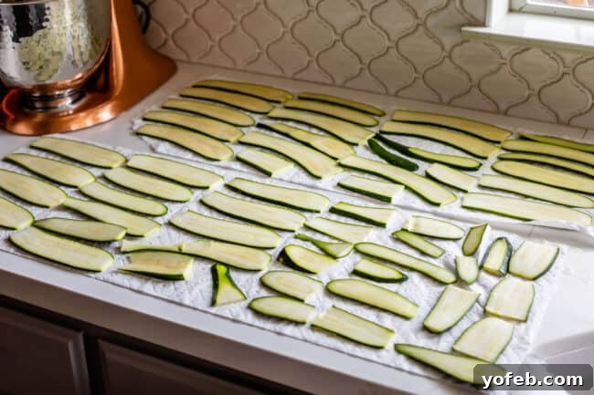 Thinly sliced zucchini strips are laid out on paper towels on a countertop, ready to absorb water through the salting method. This preparation step is crucial for making zucchini lasagna less watery. Another layer of paper towels and a generous sprinkle of kosher salt are visible, setting the stage for the moisture-wicking process.