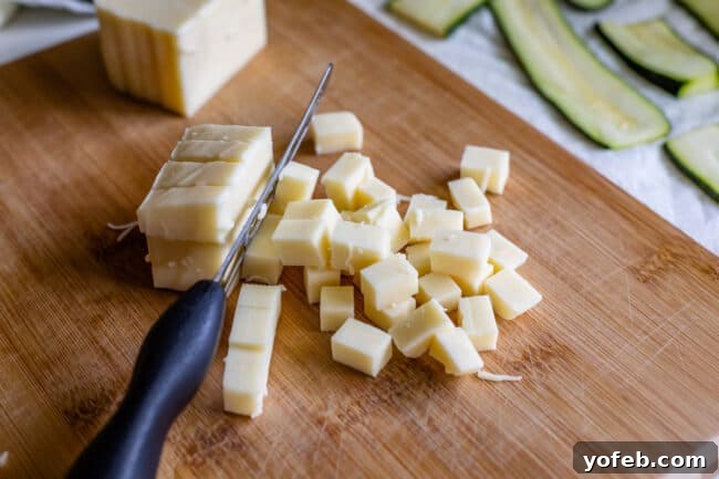 A block of mozzarella cheese is being expertly chopped into 1/2-inch cubes on a wooden cutting board with a chef's knife. This preparation method is preferred for zucchini lasagna, as cubed mozzarella melts into delightful, gooey pockets, offering a superior texture compared to shredded cheese.