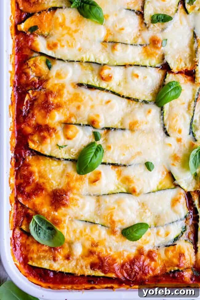 A close-up view of a baked zucchini noodle lasagna in a white baking dish, garnished with vibrant fresh basil leaves. This image showcases the rich texture and appealing layers of this low-carb, keto-friendly dish, which uses thinly sliced zucchini instead of traditional pasta, combined with Italian sausage, marinara sauce, ricotta, and mozzarella for a seriously delicious and healthy meal.