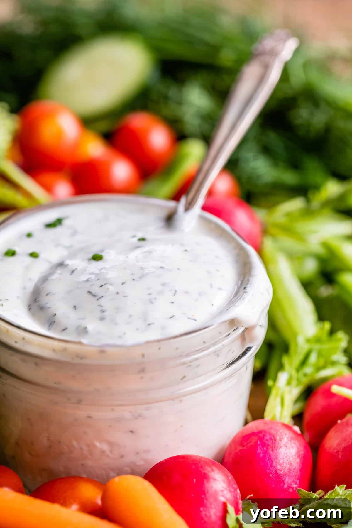 A glass jar filled with creamy buttermilk ranch dip, garnished with chopped herbs, sits surrounded by fresh vegetables, including cherry tomatoes, radishes, carrots, and celery. A spoon rests inside the jar.