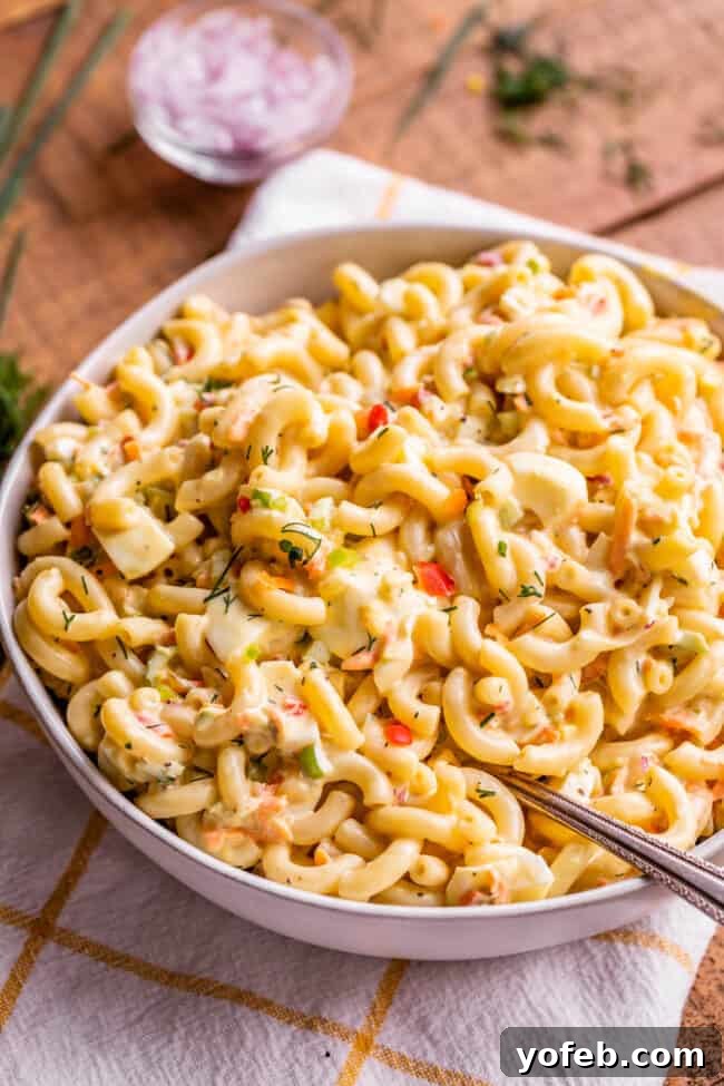 Classic Creamy Macaroni Salad 9 best macaroni salad ever in a bowl with a serving spoon, presented on a white and gold tea towel for an elegant touch.