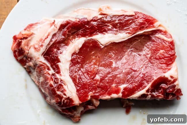 Uncooked, well-marbled ribeye steak on a simple white plate, ready for seasoning.