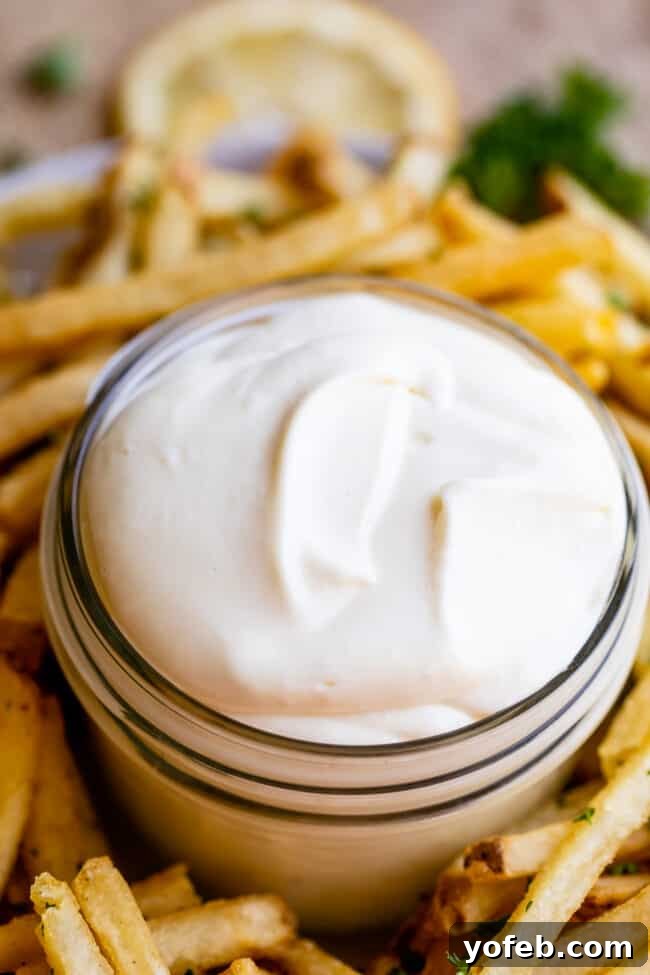 Garlic Aioli Recipe (Classic and Cheater Versions) from The Food Charlatan. I will show you just how easy it is to make homemade Garlic Aioli Sauce! Making it from scratch is quick and easy, but I have a cheater version that is even quicker and easier. Either way you make it, aioli is lemony garlicky heaven! It will be your new favorite condiment in no time. #recipe #easy #garlic #lemon #aioli #sauce #mayonnaise #roasted #garlicmayo #fromscratch #homemade #creamy A jar of garlic aioli with a blurred background of french fries, emphasizing the dip.