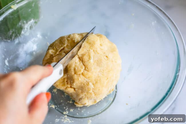 A knife cutting a ball of pie dough in half within a glass bowl, showing its readiness for chilling and shaping.