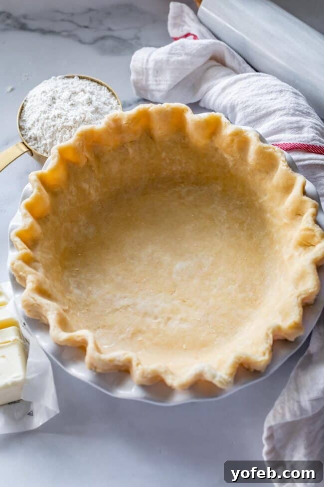 A single, easy flaky pie crust is beautifully fluted in a pan, accompanied by flour, a sliced unwrapped stick of butter, and a rolling pin, all essential tools for homemade baking.