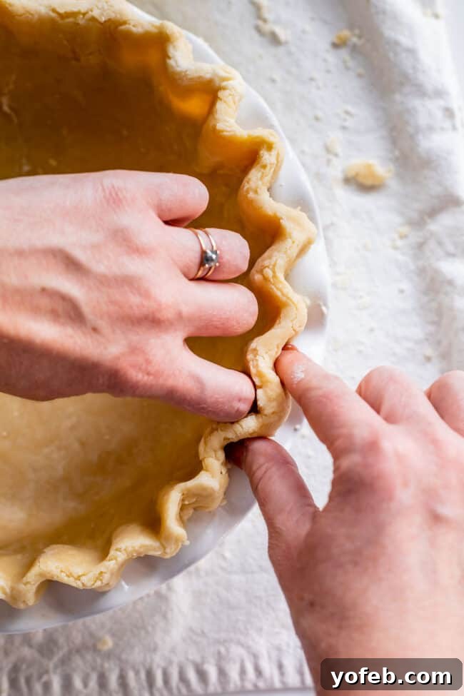 How to Pre-Bake Your Pie Crust Perfectly 6 Hands carefully fluting the edges of a raw pie crust in a ceramic dish, creating a decorative and structurally sound border before the blind baking process.