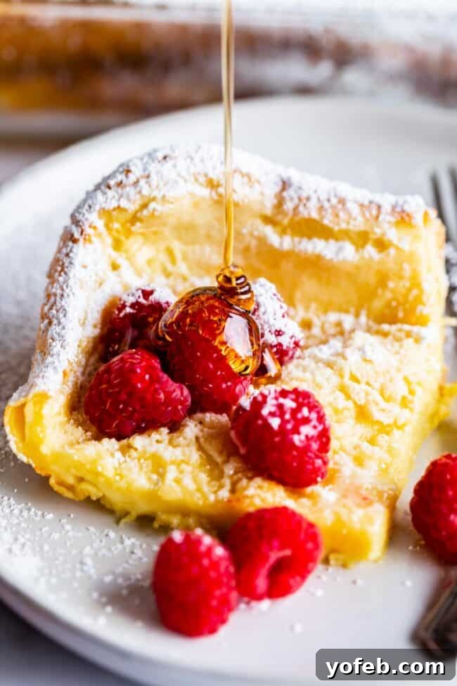 dutch baby pancake on a plate with raspberries and powdered sugar, being drizzled with syrup.