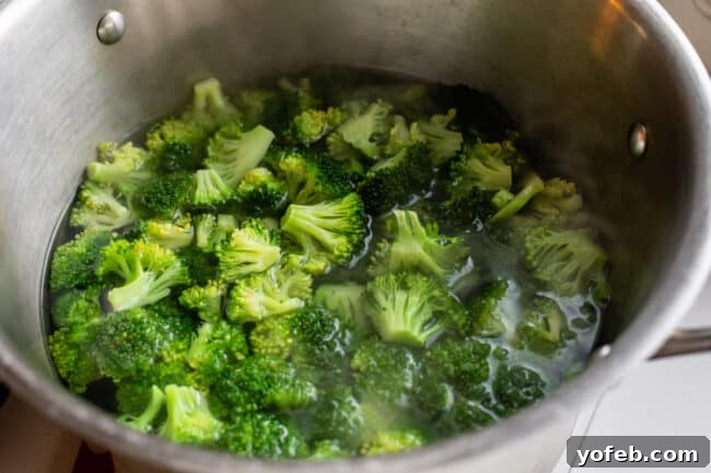 A large silver pot on a stove with fresh broccoli florets being blanched in boiling water. Steam rises from the pot, indicating the quick initial cooking step.