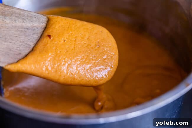 Effortless Homemade Peanut Sauce 11 A close-up view of rich Thai peanut sauce clinging to a wooden spoon, held over a saucepan, showcasing its appealing thickness and smooth texture after cooking.
