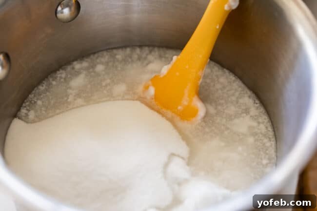 Island Oasis Piña Colada 13 A saucepan containing sugar, coconut milk, and salt, with a yellow spatula stirring the mixture, illustrating the preparation of homemade cream of coconut.
