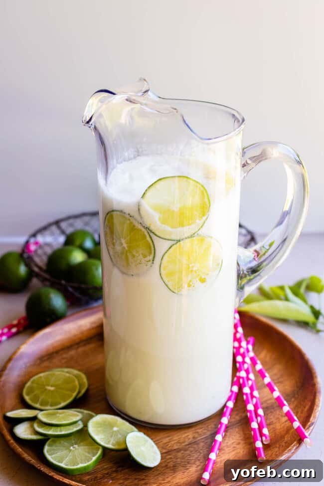 Creamy Brazilian Lemonade Recipe 17 A tall, clear glass pitcher filled with Brazilian Lemonade, complete with pink polka dot straws and lime slices for serving.