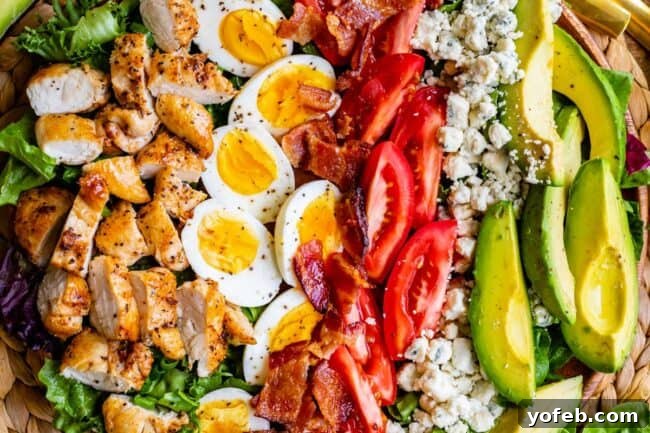 A classic Cobb salad arranged in neat rows within a clear glass bowl, showcasing the distinct layers of chicken, bacon, cheese, eggs, tomatoes, avocado, and lettuce.
