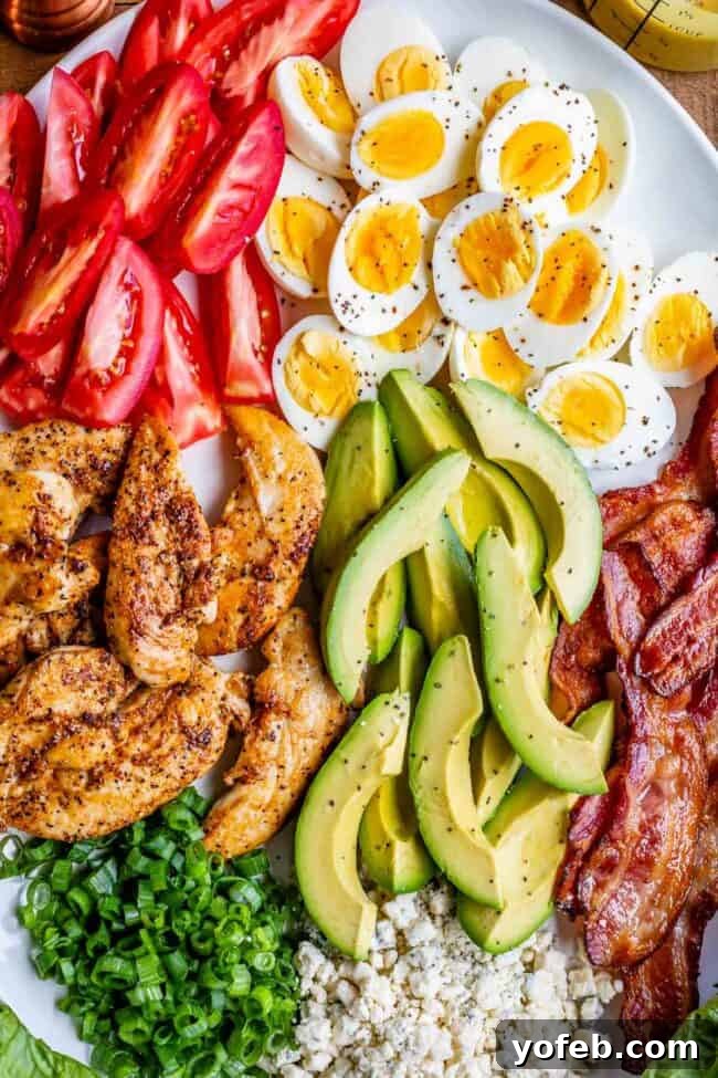 Assortment of fresh Cobb salad ingredients, including chicken, bacon, hard-boiled eggs, and various chopped vegetables, neatly arranged on a pristine white serving platter.