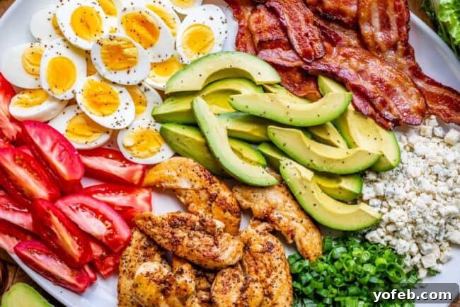All the essential components of a traditional Cobb salad, including neatly arranged piles of grilled chicken, crumbled bacon, hard-boiled eggs, diced tomatoes, fresh avocado, green onions, and blue cheese, displayed on a clean white platter.