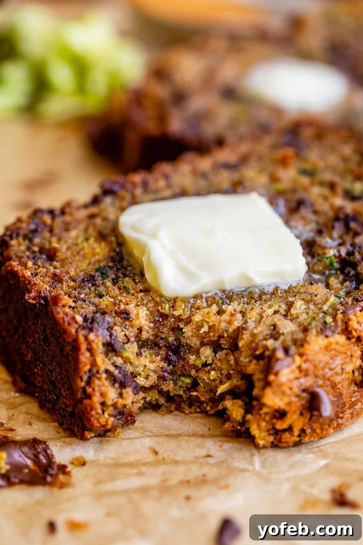 Heavenly Zucchini Bread 28 A close-up of zucchini bread with chocolate chips, topped with a pat of melting butter, rests on parchment paper, with another slice blurred in the background.