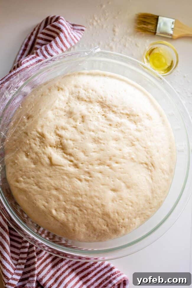 Foolproof Pizza Dough for Newbies 18 pizza dough rising in a glass bowl with plastic wrap.