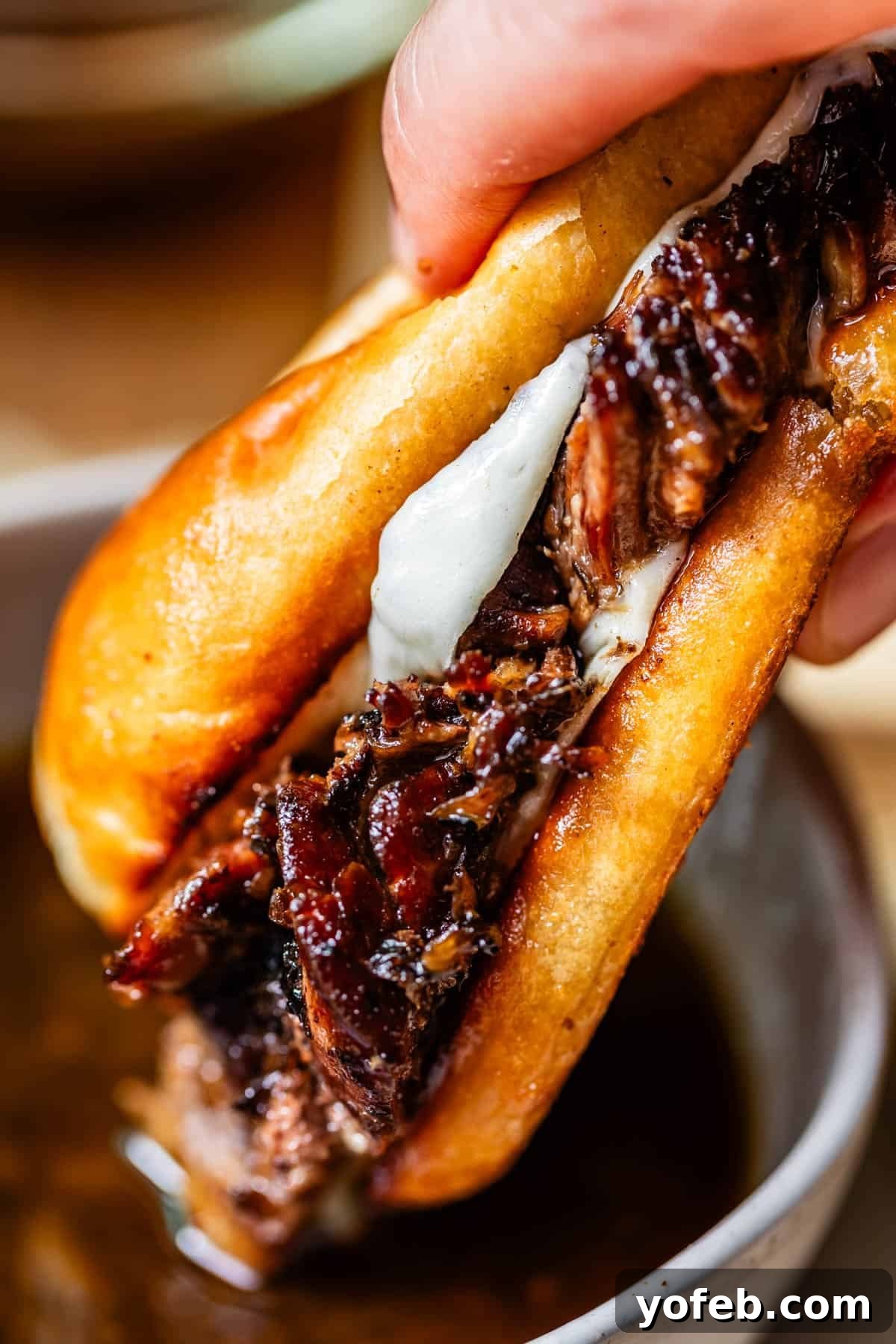 Ultimate Slow Cooker French Dip Sandwiches 2 A close-up of a hand holding a French dip sandwich filled with juicy, shredded beef and melted cheese, with a bowl of au jus dipping sauce underneath. Perfect for comfort food enthusiasts.