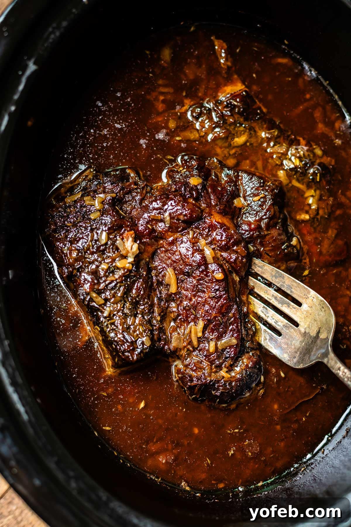 Ultimate Slow Cooker French Dip Sandwiches 14 A close-up of a perfectly cooked beef roast in a slow cooker, submerged in a dark, rich, and aromatic sauce, with visible seasonings and herbs. A metal fork gently rests on the incredibly tender, juicy meat, indicating its readiness to be shredded or sliced.