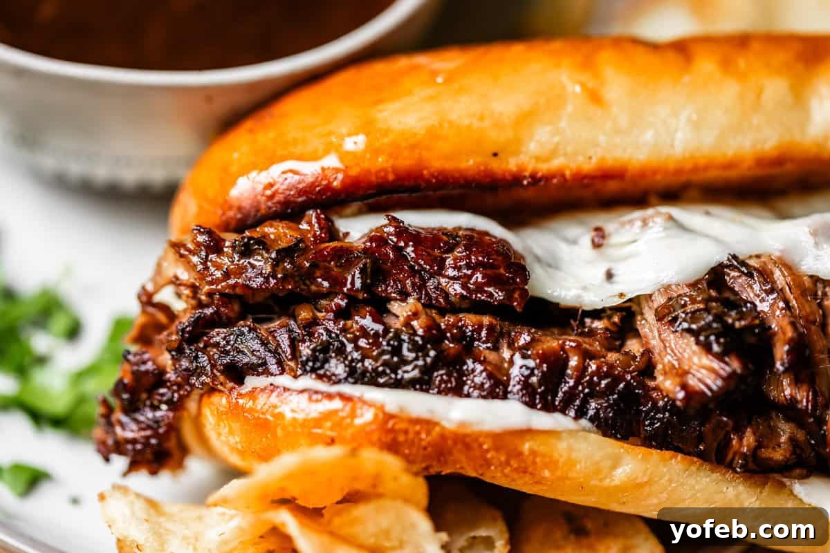 Ultimate Slow Cooker French Dip Sandwiches 20 Close-up of a perfectly constructed French dip sandwich with shredded, juicy beef and melted cheese on a toasted bun, served with a side of crispy potato chips and a cup of rich au jus dipping sauce blurred in the background, ready for enjoyment.