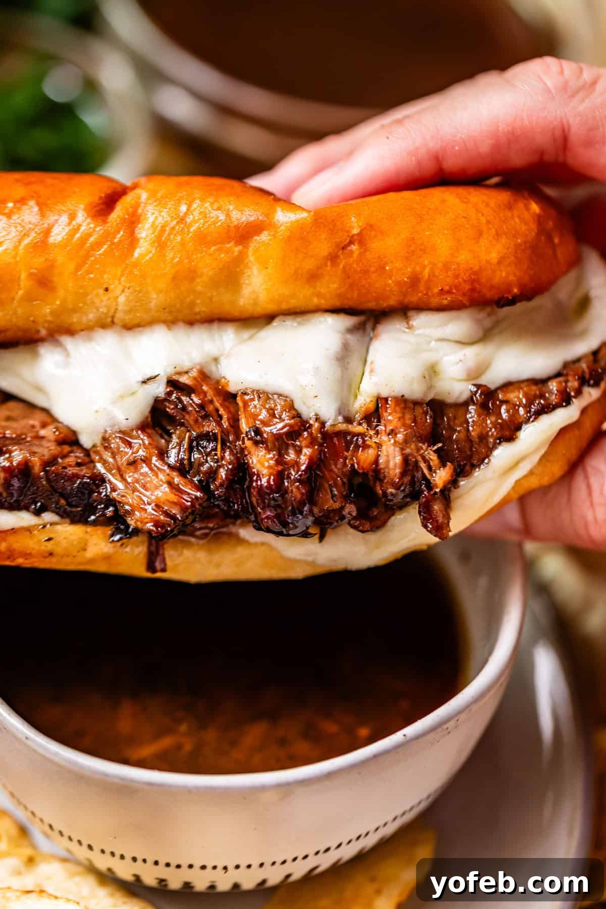 Ultimate Slow Cooker French Dip Sandwiches 3 A hand holds a sandwich filled with shredded roast beef and melted cheese on a toasted bun, with a small bowl of au jus dipping sauce underneath, highlighting the perfect bite.
