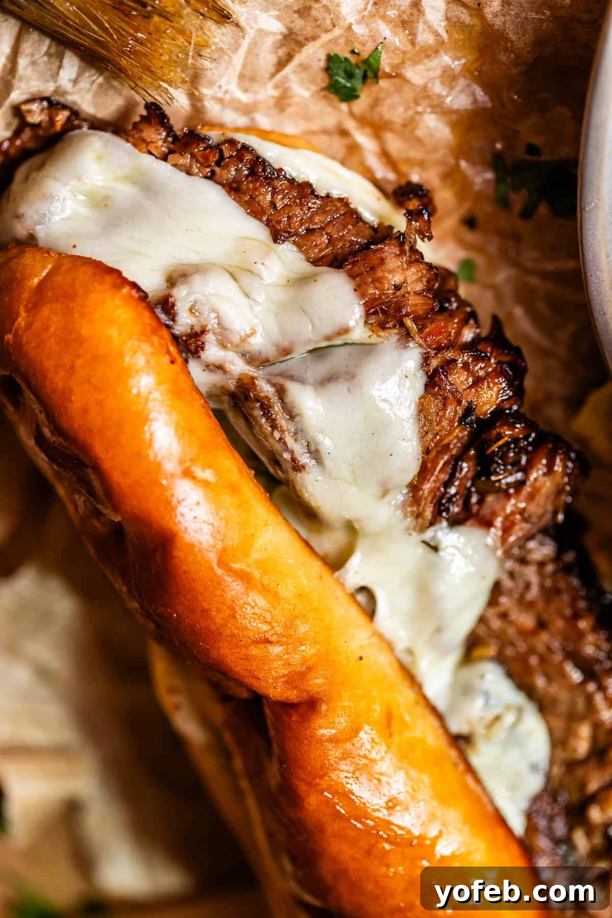 Ultimate Slow Cooker French Dip Sandwiches 21 Close-up of a toasted sandwich roll filled with perfectly grilled steak and generously melted cheese, artfully resting on brown parchment paper with fresh parsley sprinkled around, enhancing its visual appeal.