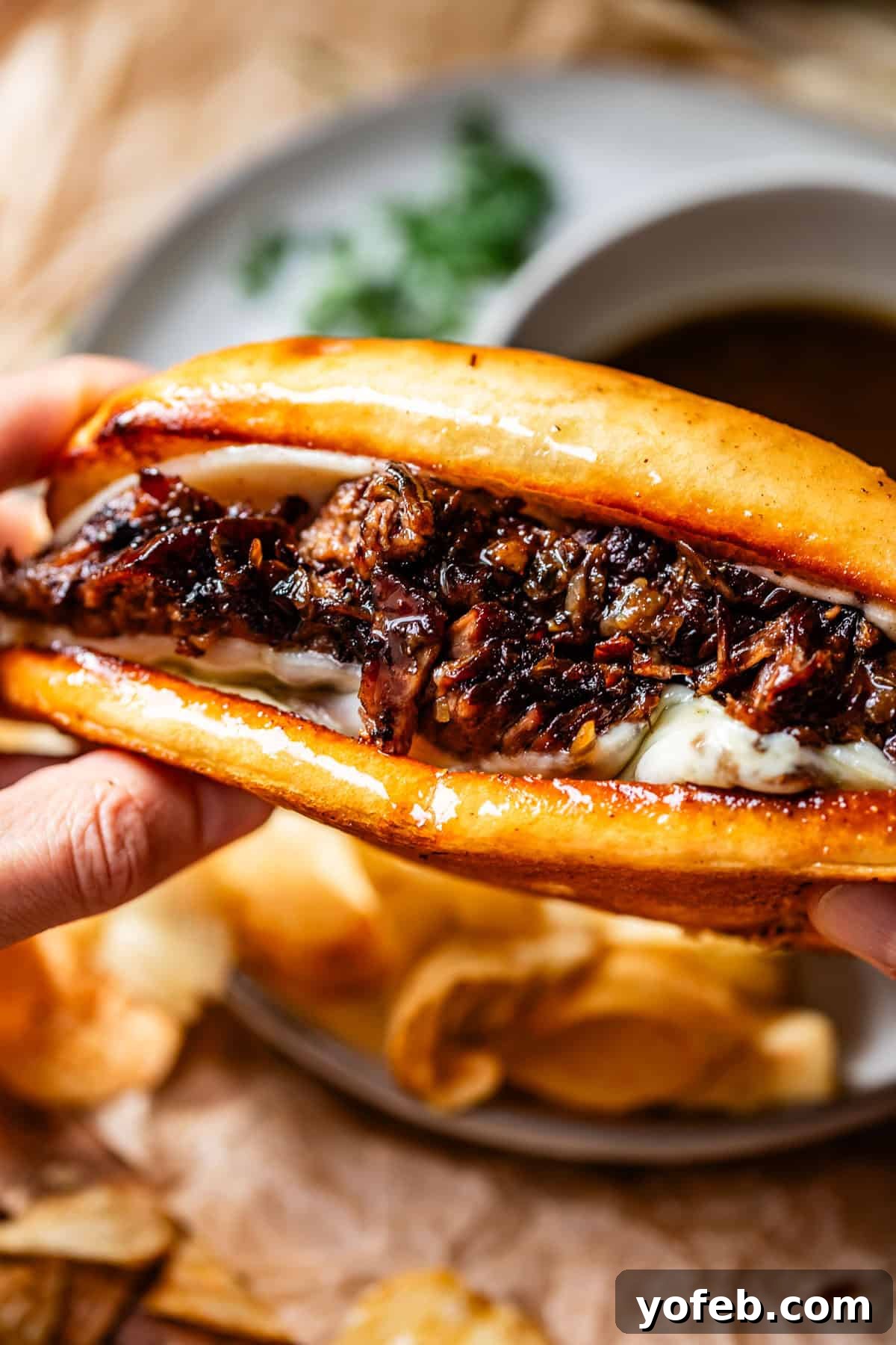 Ultimate Slow Cooker French Dip Sandwiches 22 A close-up of hands holding a French dip sandwich filled with tender, sliced beef and melted cheese, with a side of crispy potato chips and a bowl of steaming au jus in the background, ready for dipping.
