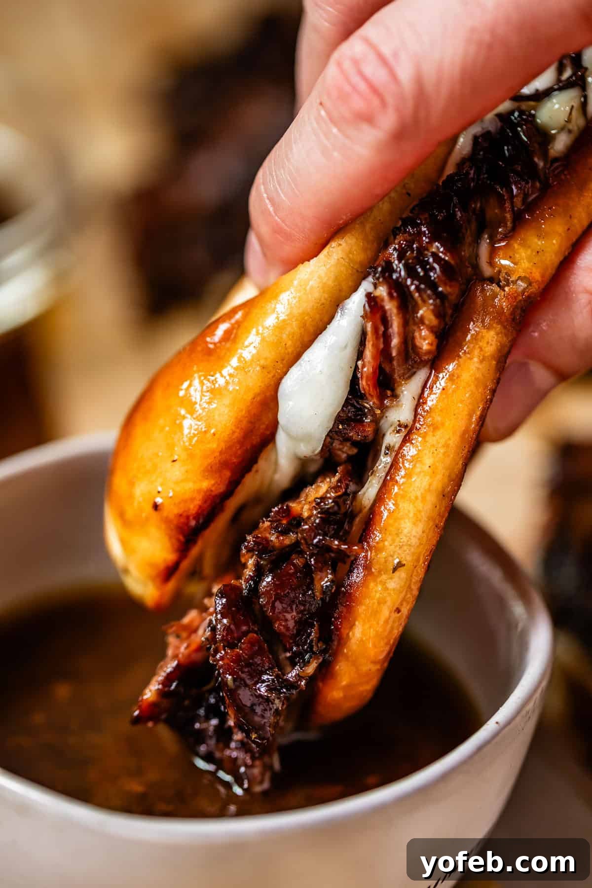 Ultimate Slow Cooker French Dip Sandwiches 24 A hand dips a juicy, grilled beef sandwich with melted cheese into a small bowl of rich, brown au jus sauce. The sandwich is packed with tender meat and served on a golden, perfectly toasted bun, ready for a delicious bite.