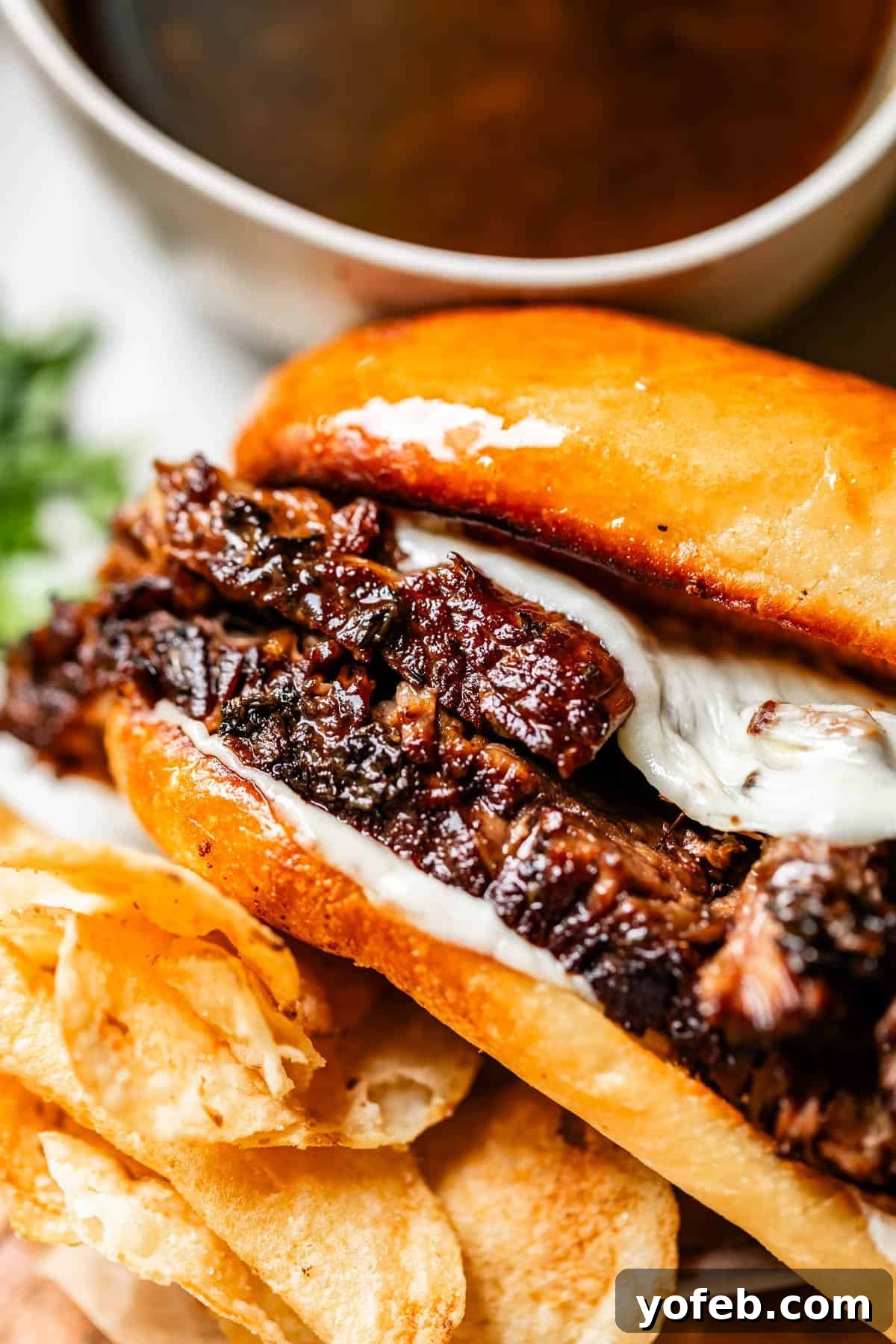 Ultimate Slow Cooker French Dip Sandwiches 4 A close-up of a French dip sandwich with tender beef and melted cheese in a toasted bun, served with crispy potato chips and a steaming cup of au jus dipping sauce in the background.