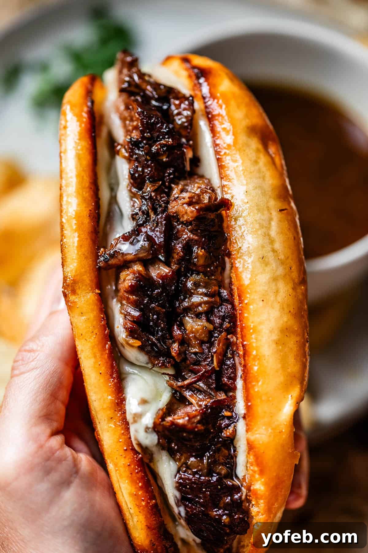 Ultimate Slow Cooker French Dip Sandwiches 5 A close-up of a hand holding a toasted sandwich filled with melted cheese and tender, juicy pieces of beef, with a cup of au jus sauce blurred in the background, ready for dipping.