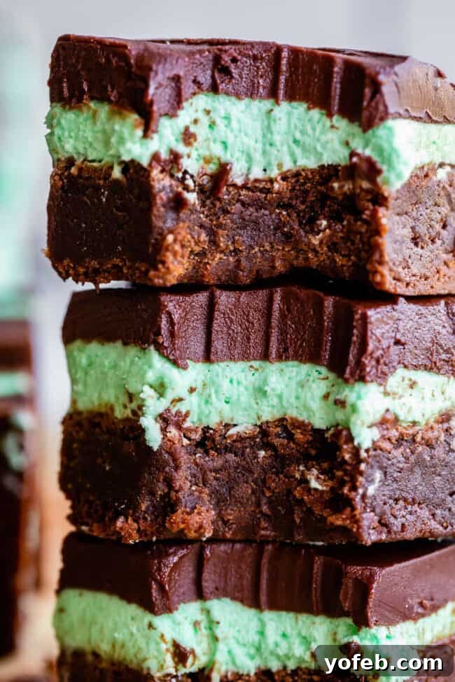 A close-up of three stacked mint brownies, each with a fudgy chocolate base, a green mint cream layer, and a thick chocolate topping. The top brownie has a bite taken out of it.