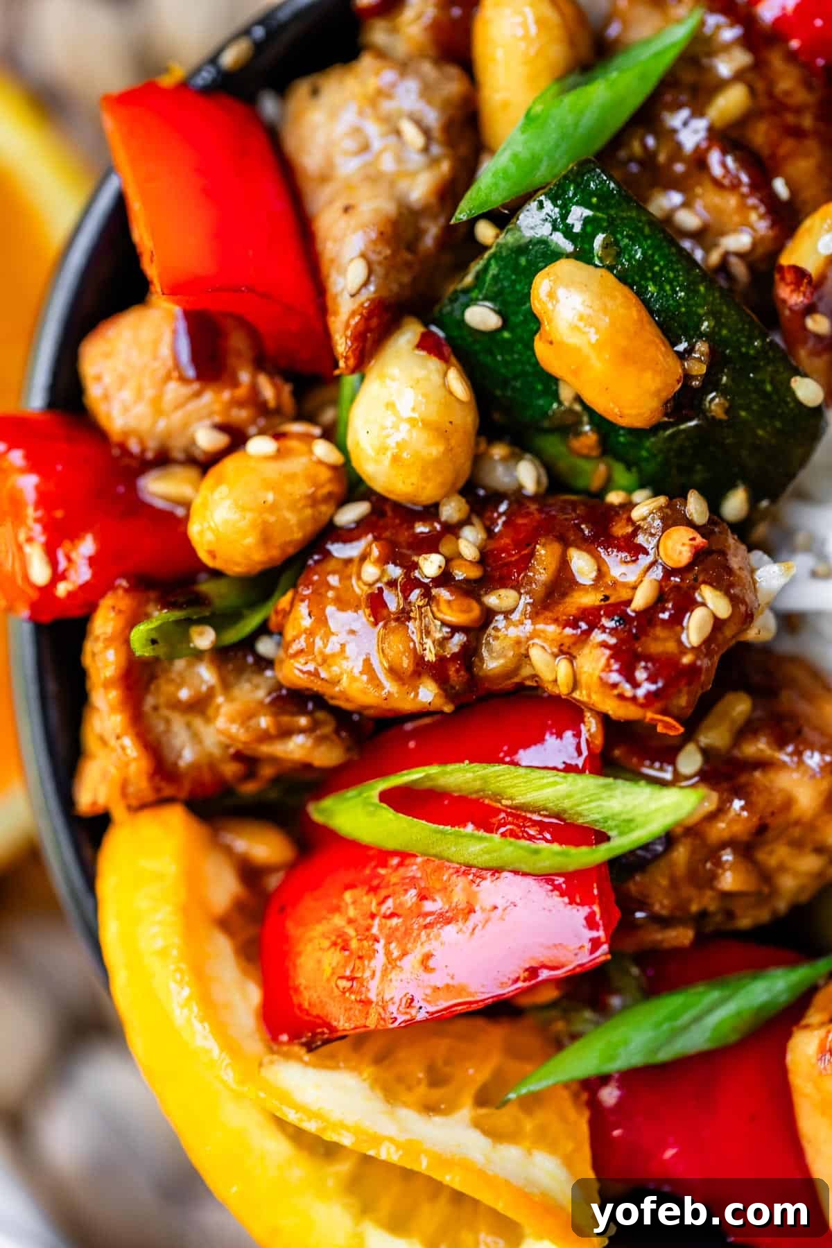 Szechuan Kung Pao Chicken 5 Close-up of Kung Pao chicken in a sleek black ceramic bowl, adorned with bright red and green vegetables, chicken pieces, and a generous sprinkle of sesame seeds, all coated in a rich, savory sauce.