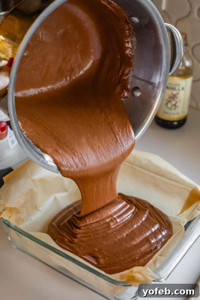 Mom's Effortless Fudge 24 Smooth, glistening fudge being poured from a pot into a glass pan meticulously lined with parchment paper.