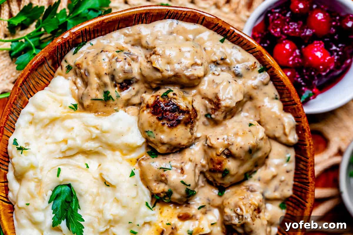 Grandma Prudy's Timeless Swedish Meatballs 22 A beautiful ceramic bowl filled with a generous serving of mashed potatoes, topped with tender Swedish Meatballs and a rich brown sauce.
