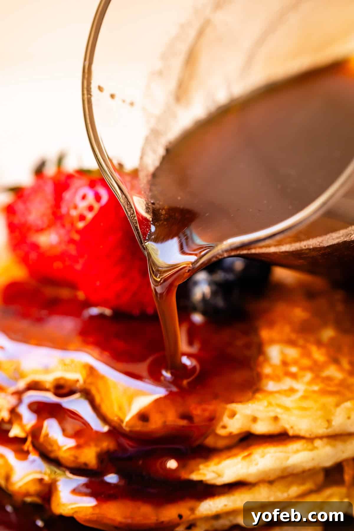 Homemade maple syrup being poured out of a glass spout over pancakes with strawberries in the background.