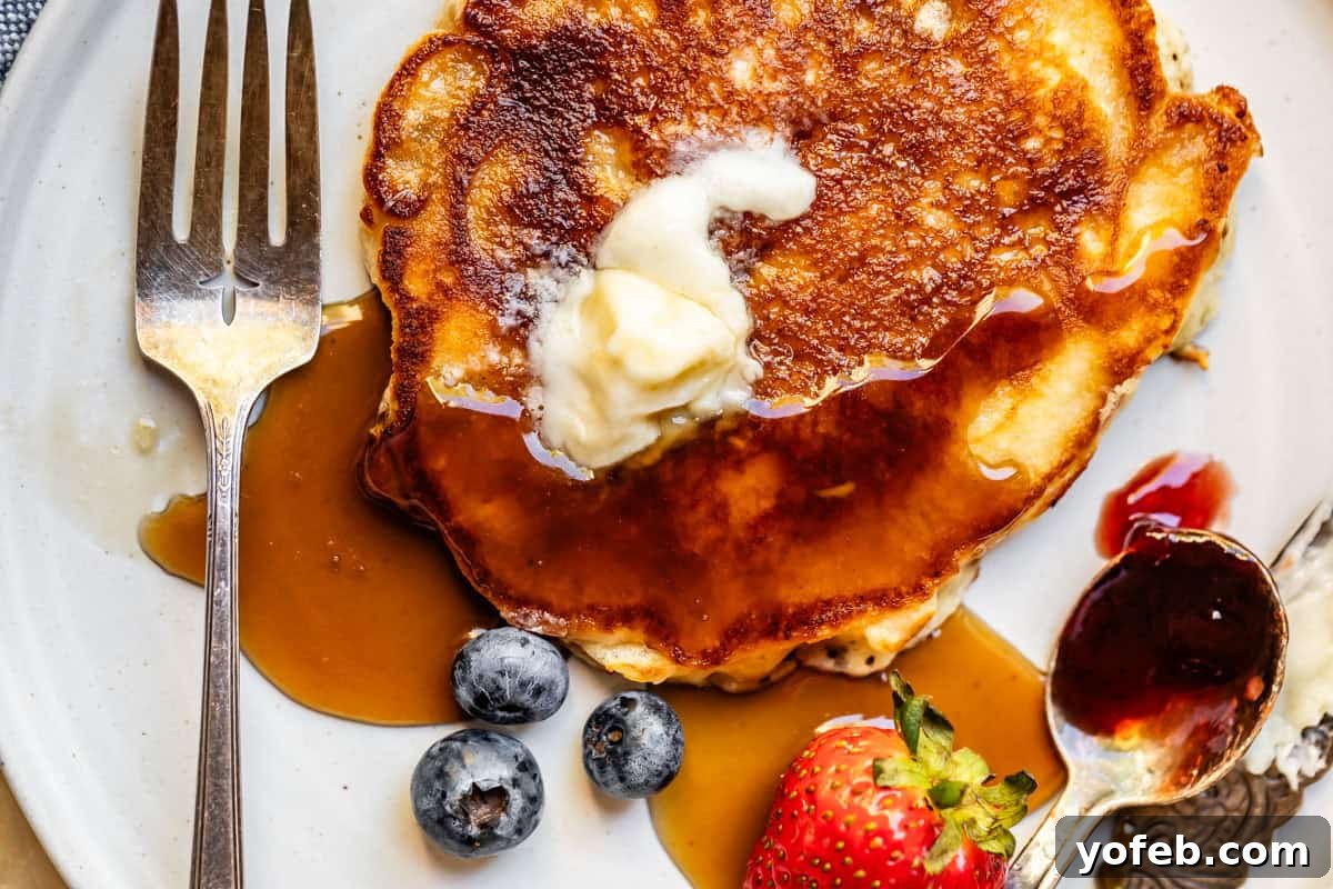 Effortless Homemade Maple Syrup 9 A beautifully browned pancake on a plate, adorned with a pat of melting butter and generous drizzles of homemade syrup, accompanied by a fork and knife, and fresh fruit.