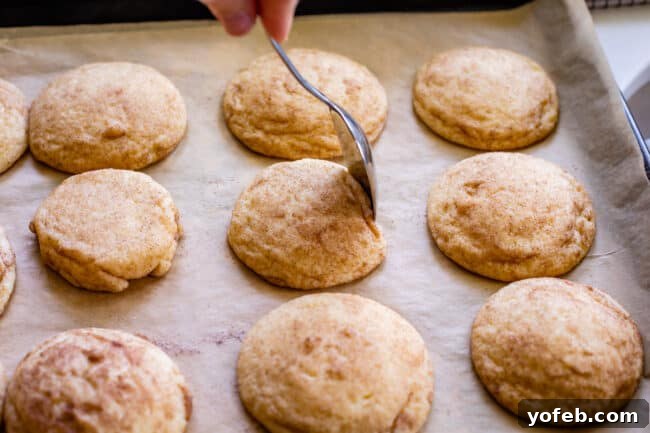 A spoon gently shaping the edge of a warm Snickerdoodle cookie on a baking sheet immediately after baking, pushing it towards the center.