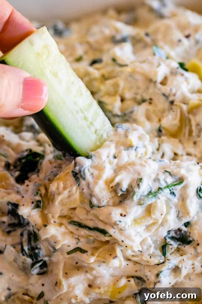 Ultimate Cheesy Spinach Artichoke Dip 17 Hand elegantly dipping a fresh cucumber spear into a creamy, healthy spinach and artichoke dip.
