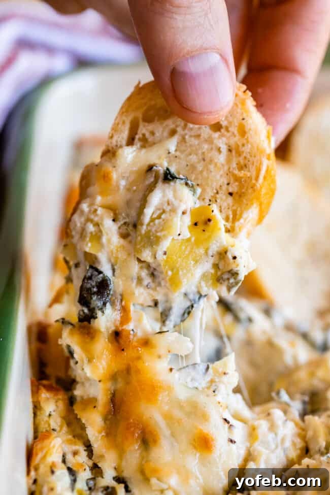 Ultimate Cheesy Spinach Artichoke Dip 19 Hand dipping a golden toasted bread slice into a bubbling hot spinach artichoke dip, revealing an enticing cheese pull.