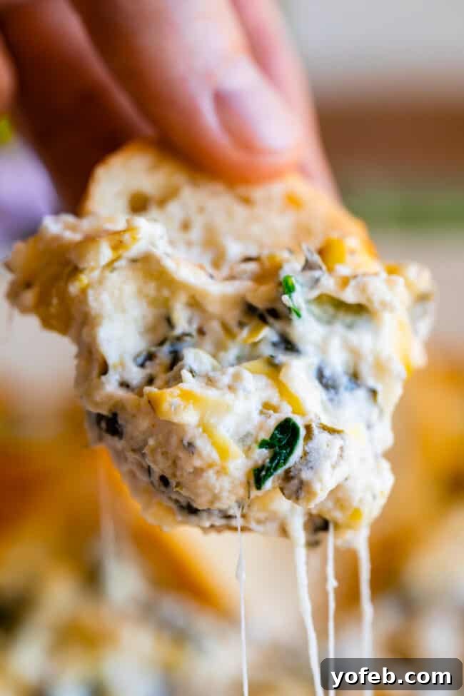 Ultimate Cheesy Spinach Artichoke Dip 6 Hand holding a slice of bread expertly dipped into a warm spinach artichoke dip, showcasing a perfect cheese pull.
