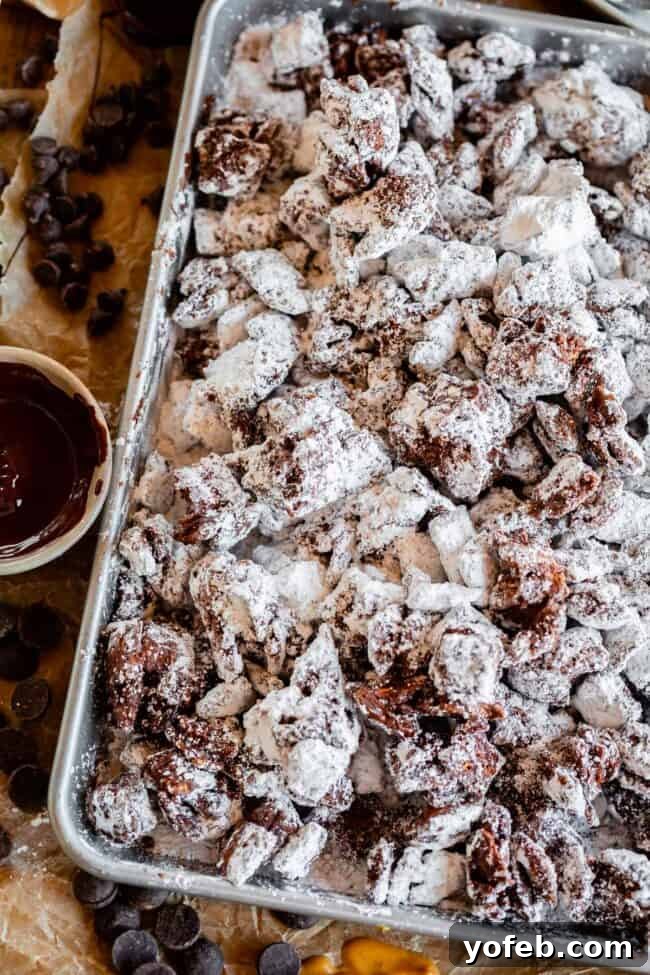 Loaded Chunky Chex Muddy Buddies 4 Muddy Buddies cooling on a sheet pan, with a bowl of melted chocolate and peanut butter in the foreground.