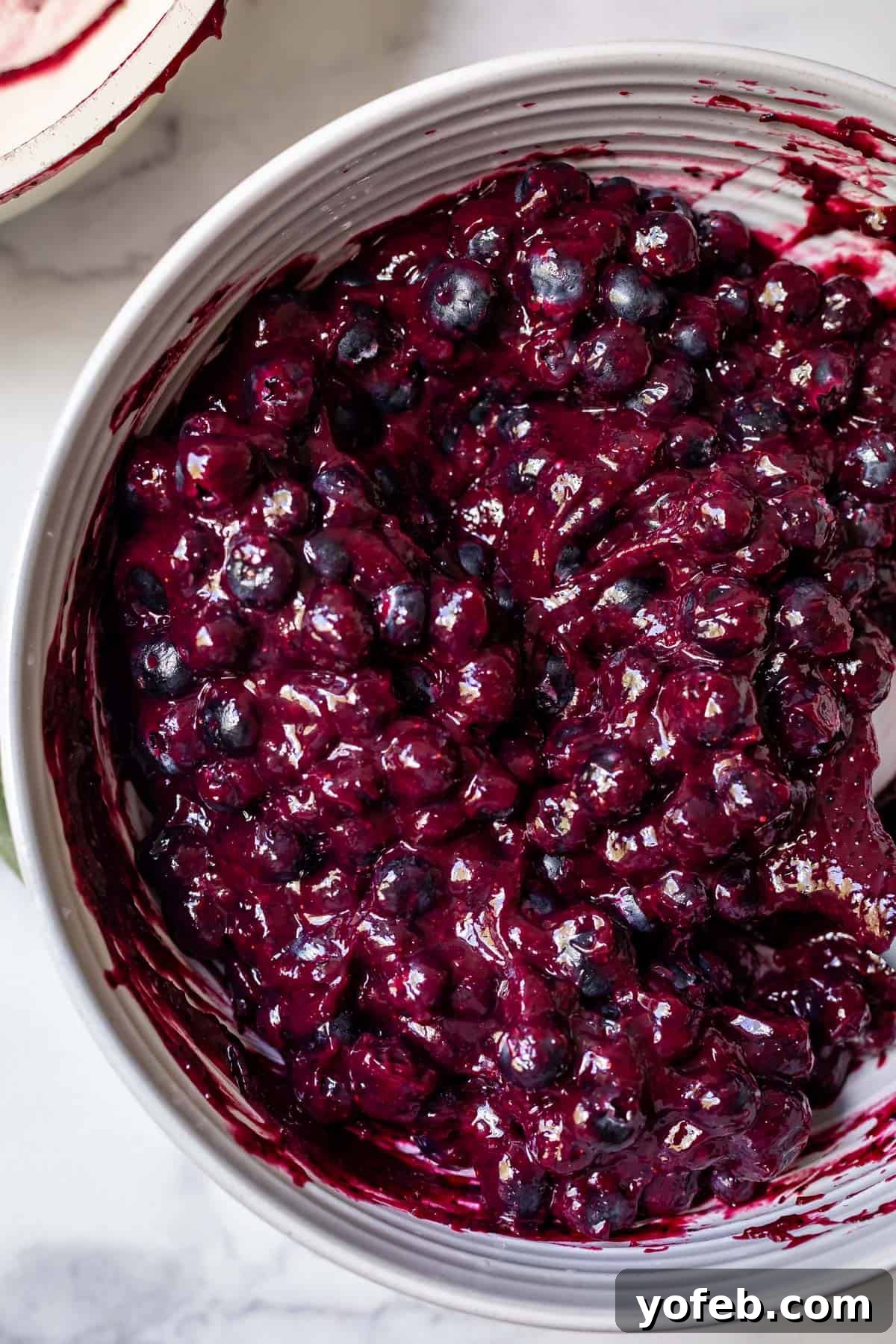 Grandma's Classic Blueberry Pie 17 An overhead shot of a white ceramic bowl filled with glossy, dark purple blueberry pie filling, showcasing the mixture of whole and puréed berries.