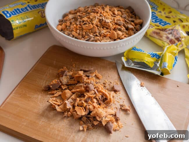 The Ultimate Peanut Butter Butterfinger Cookie 14 A bowl filled with Butterfinger Baking Bits sits in the background, while in the foreground, freshly chopped Butterfinger candy bars are displayed on a cutting board, illustrating that both forms of the candy work equally well for this recipe.