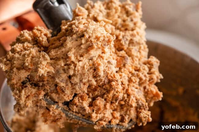 The Ultimate Peanut Butter Butterfinger Cookie 16 A close-up shot of rich peanut butter Butterfinger cookie dough clinging to the paddle attachment of a stand mixer, showcasing the abundance of crunchy candy bits mixed throughout.