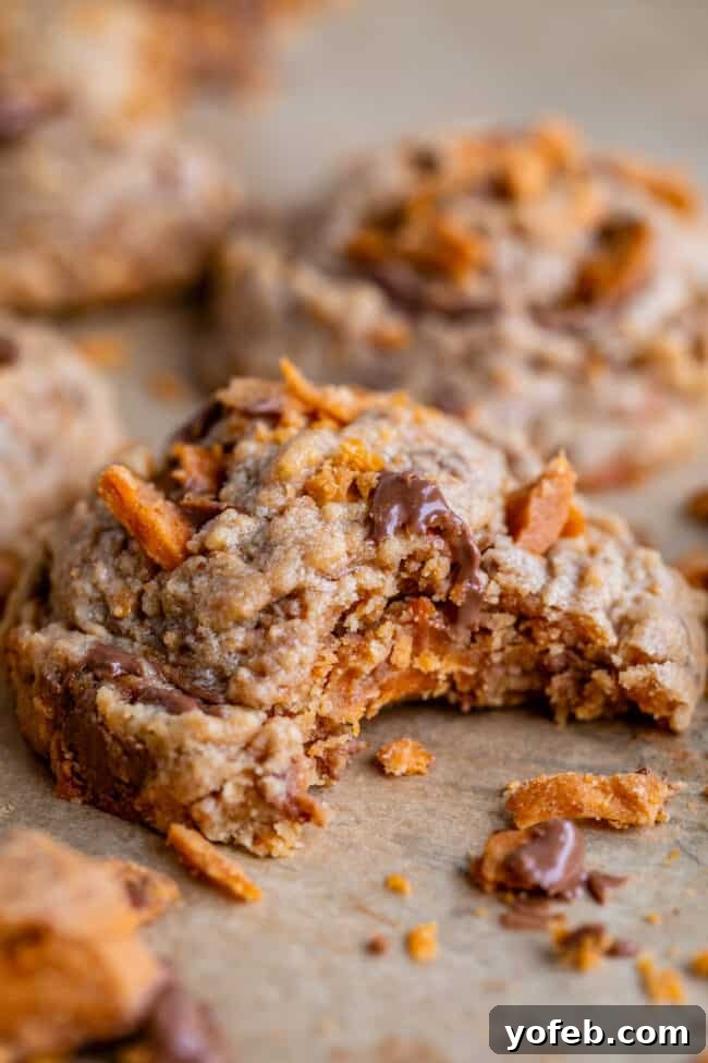 The Ultimate Peanut Butter Butterfinger Cookie 20 A close-up side profile of a delicious Peanut Butter Butterfinger Cookie with a bite taken out, resting on parchment paper. The image highlights the cookie's thick, soft texture and the crunchy Butterfinger pieces embedded within.