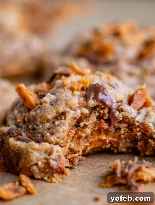 The Ultimate Peanut Butter Butterfinger Cookie 3 Close-up side view of a Peanut Butter Butterfinger Cookie with a bite taken out, revealing its soft interior and crunchy Butterfinger pieces. The cookie sits on parchment paper, emphasizing its homemade appeal.