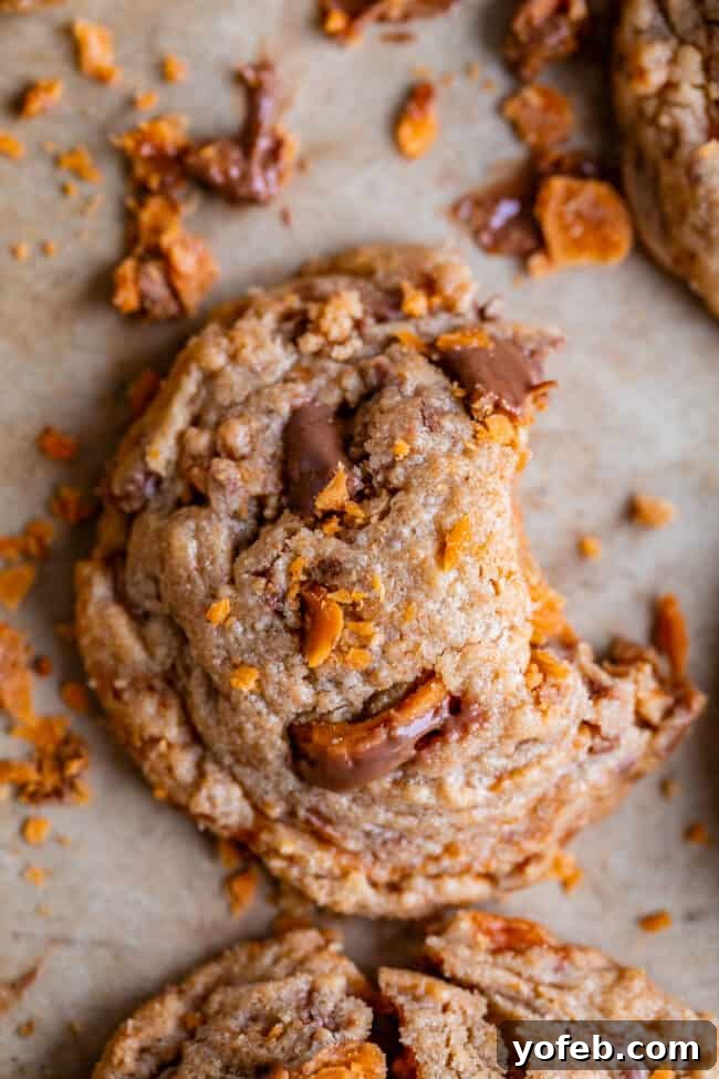 The Ultimate Peanut Butter Butterfinger Cookie 7 An overhead shot of a freshly baked Butterfinger cookie with a small bite taken out, showcasing the melted candy pieces and the soft, golden-brown cookie base. The cookie is presented on a parchment-lined baking sheet, emphasizing its homemade quality.